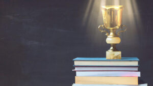 Golden trophy on pile of books, against blackboard, with sun rays over trophy; learning/achievement concept