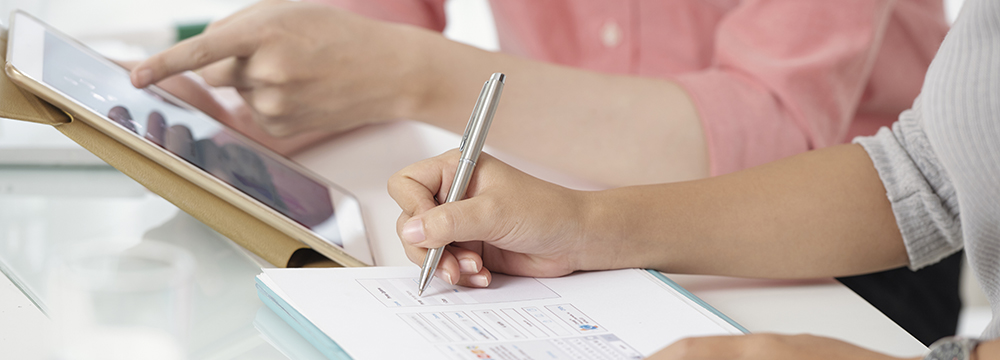 Faceless shot of contemporary colleagues using tablet while writing on clipboard sitting at table