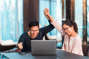 Young Asian married couple hold hands celebrating together, watching laptop computer in modern home office. Success job, love relationship, football sport cheering, or small business startup concept.